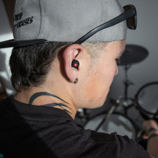 Músico con gorra usando los tapones para los oídos reutilizables Mood Earplugs Happy Black mientras toca la batería